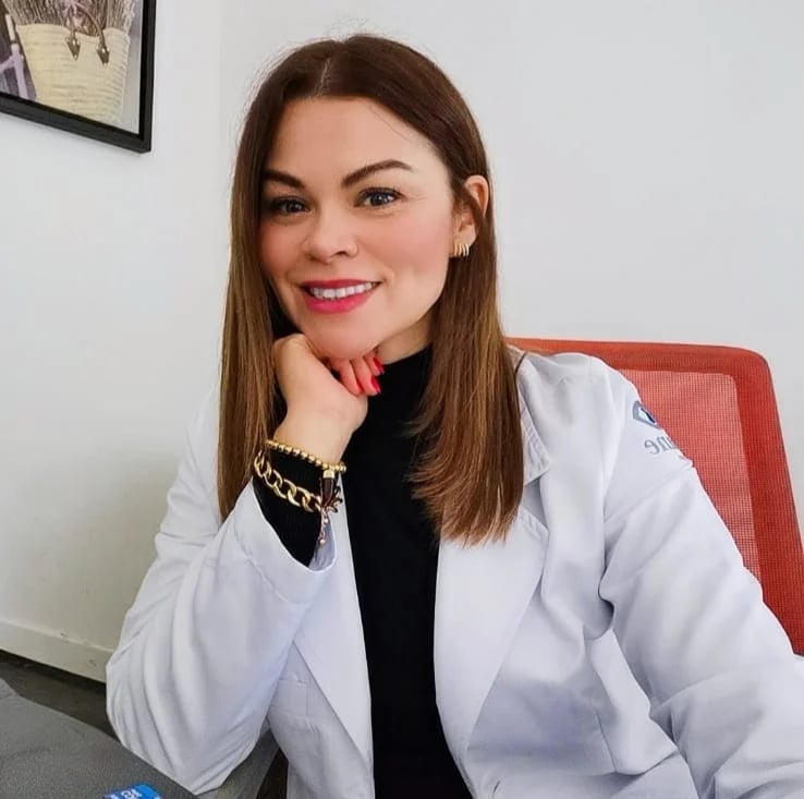 Mujer sonriente con bata médica blanca y camisa negra sentada en oficina